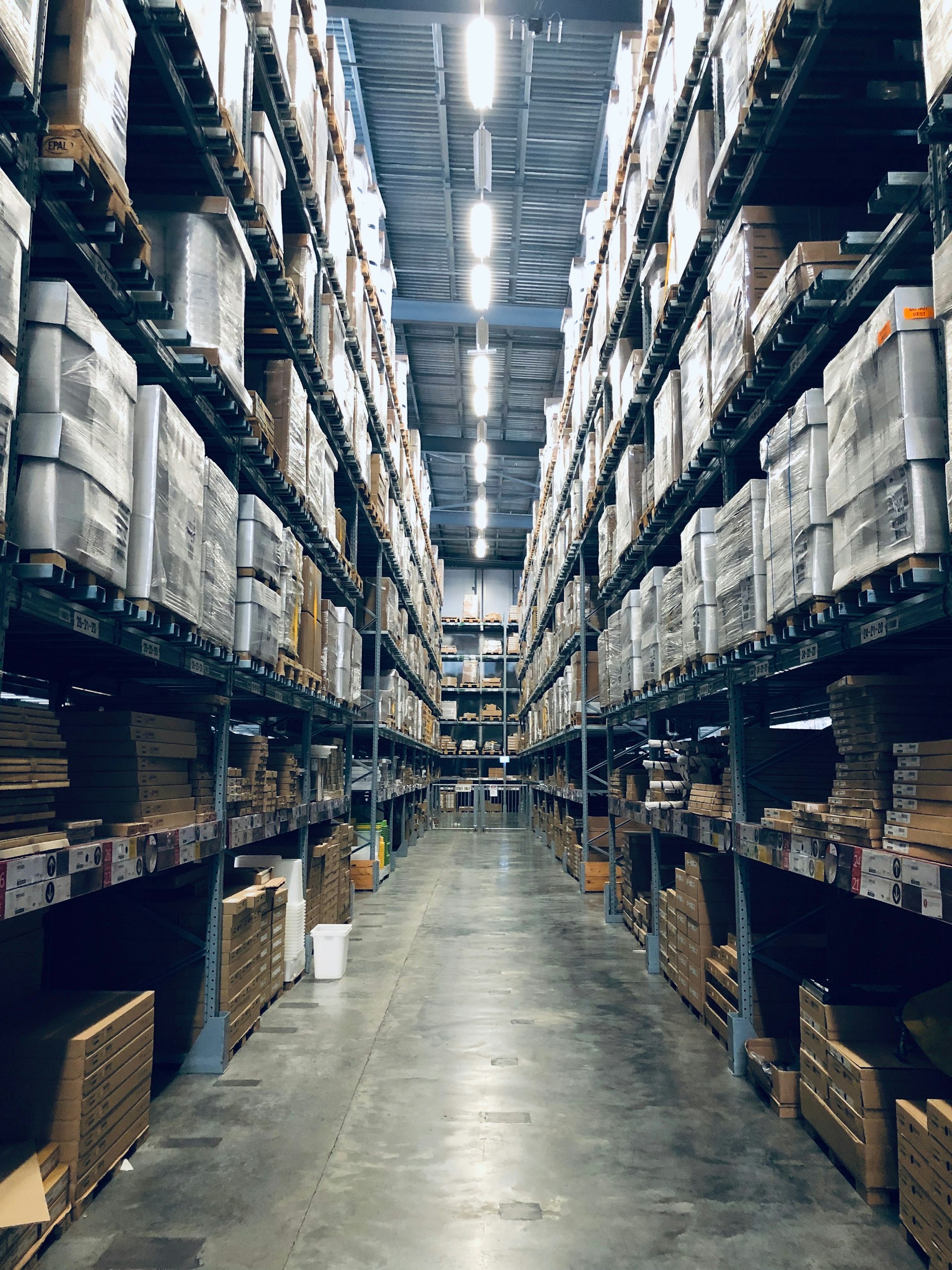 Tall warehouse aisle stacked with palletised inventory under bright overhead lighting